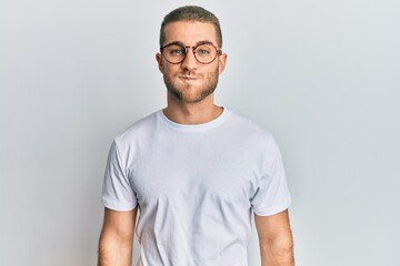 Young caucasian man wearing casual clothes and glasses puffing cheeks with funny face. mouth inflated with air, crazy expression.