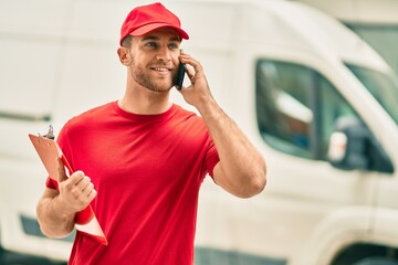 Young caucasian deliveryman talking on the smartphone and holding clipboard at the city.