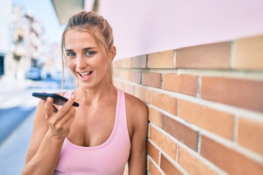 Young blonde sporty girl talking on the smartphone leaning on the wall at street of city.