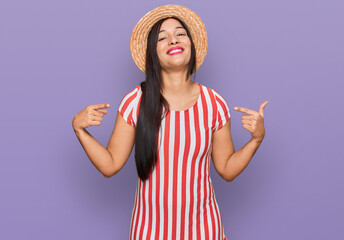 Young hispanic woman wearing summer hat looking confident with smile on face, pointing oneself with...