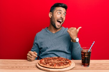 Handsome hispanic man eating tasty pepperoni pizza pointing thumb up to the side smiling happy with open mouth