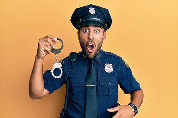Handsome hispanic man wearing police uniform holding metal handcuffs scared and amazed with open mouth for surprise, disbelief face