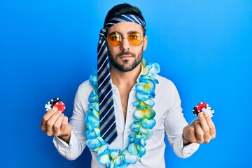 Young hispanic businessman wearing party funny style holding casino chips relaxed with serious...
