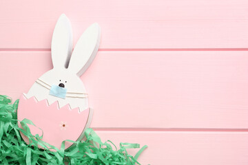 Cute bunny figure in protective mask on pink wooden background, top view with space for text. Easter holiday during COVID-19 quarantine