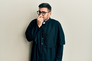 Young hispanic man wearing priest uniform standing over white background smelling something stinky...