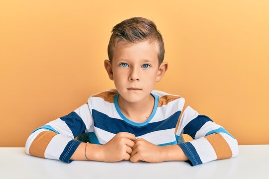 Adorable caucasian kid wearing casual clothes sitting on the table relaxed with serious expression on face. simple and natural looking at the camera.