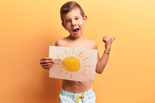 Cute blond kid wearing swimwear and summer hat pointing thumb up to the side smiling happy with open mouth