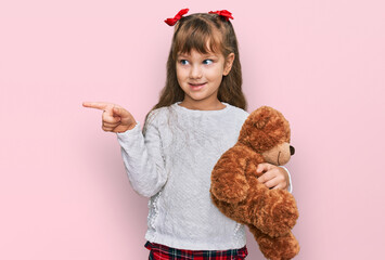 Little caucasian girl kid hugging teddy bear stuffed animal smiling happy pointing with hand and...