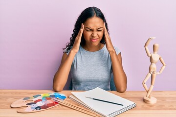 Young african american girl artist sitting at studio table with hand on head, headache because...