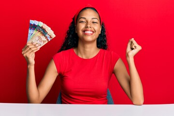 Young african american girl holding canadian dollars sitting on the table screaming proud,...