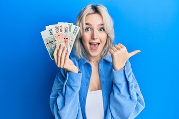 Young blonde girl holding czech koruna banknotes pointing thumb up to the side smiling happy with open mouth