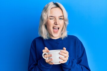 Young blonde girl drinking a cup of coffee winking looking at the camera with sexy expression, cheerful and happy face.