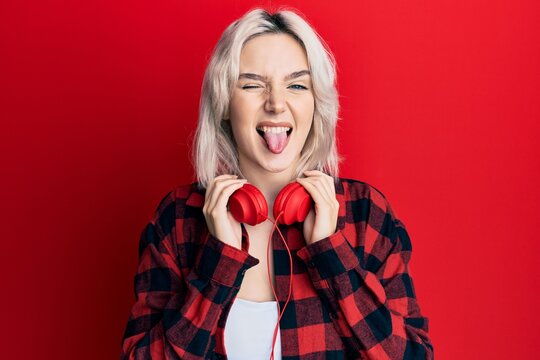 Young blonde girl listening to music using headphones sticking tongue out happy with funny expression.