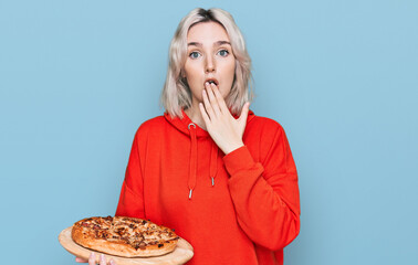 Young blonde girl holding italian pizza covering mouth with hand, shocked and afraid for mistake....