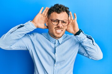 Young hispanic man wearing casual clothes and glasses trying to hear both hands on ear gesture, curious for gossip. hearing problem, deaf