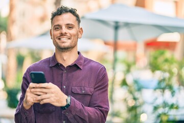 Young hispanic man smiling happy using smartphone at the city.