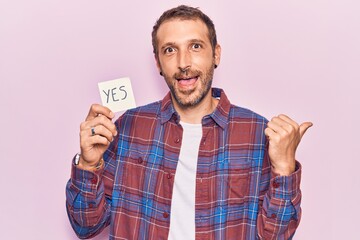 Young handsome man holding yes reminder pointing thumb up to the side smiling happy with open mouth