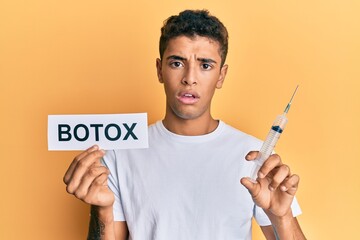 Young handsome african american man holding botox banner and needle clueless and confused...