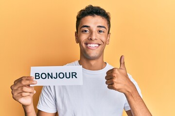 Young handsome african american man holding bonjour french greeting word smiling happy and positive, thumb up doing excellent and approval sign © Krakenimages.com