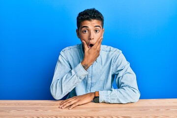 Young handsome african american man wearing casual clothes sitting on the table looking fascinated with disbelief, surprise and amazed expression with hands on chin