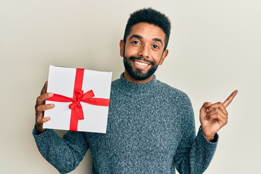 Handsome hispanic man with beard holding gift smiling happy pointing with hand and finger to the side