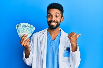 Handsome hispanic man with beard wearing doctor uniform holding 100 brazilian reals pointing thumb...