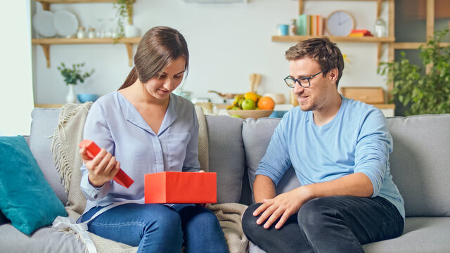 Ordering Gifts Online. A Man Gives A Woman A Gift That He Ordered Through The Site, An Online Store. The Girl Takes Out A Red Gift From A Cardboard Box. Happy Family Time. Enjoy Each Other.