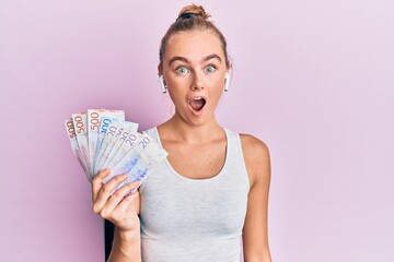 Beautiful blonde sport woman holding 20 swedish krona banknotes scared and amazed with open mouth for surprise, disbelief face