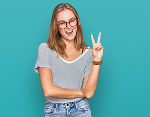 Beautiful young blonde woman wearing casual clothes and glasses smiling with happy face winking at the camera doing victory sign. number two.