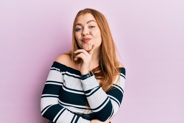 Young beautiful redhead woman wearing striped sweater over pink background smiling looking confident at the camera with crossed arms and hand on chin. thinking positive.