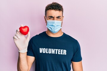 Hispanic young man wearing medical mask wearing volunteer t shirt holding heart skeptic and...