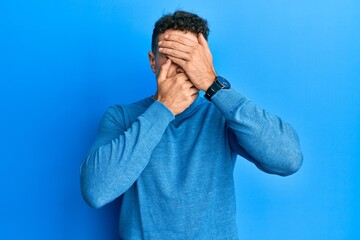Hispanic young man wearing casual winter sweater covering eyes and mouth with hands, surprised and shocked. hiding emotion