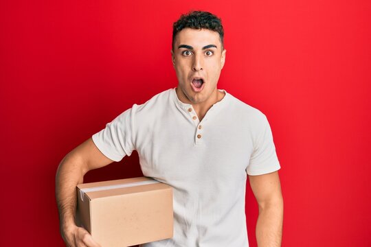 Hispanic young man holding delivery package scared and amazed with open mouth for surprise, disbelief face