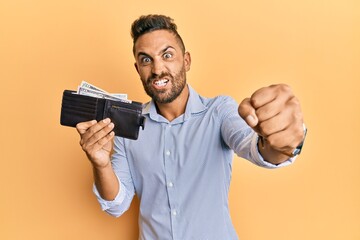 Handsome man with beard holding wallet with dollars annoyed and frustrated shouting with anger, yelling crazy with anger and hand raised