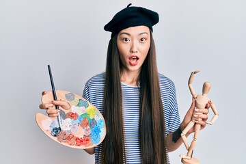 Young chinese woman wearing artist look with beret holding manikin afraid and shocked with surprise and amazed expression, fear and excited face.