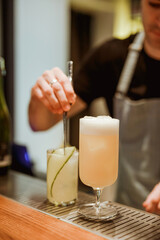 Bartender preparing gin and tonic with cucumber in the background, pina colada cocktail in the foreground. Photo with shallow depth of field. Vertical lifestyle image.