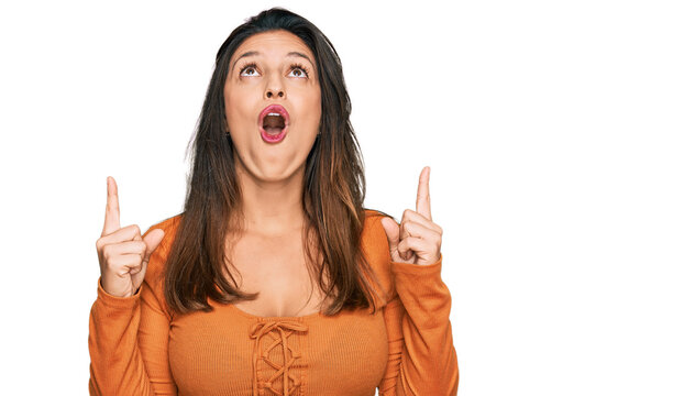 Beautiful hispanic woman wearing casual clothes amazed and surprised looking up and pointing with fingers and raised arms.