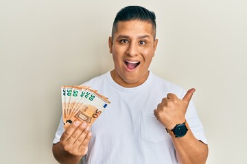 Young latin man holding bunch of 50 euro banknotes pointing thumb up to the side smiling happy with open mouth