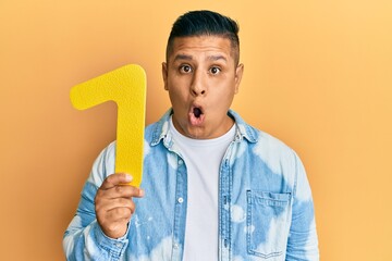 Young latin man holding number one scared and amazed with open mouth for surprise, disbelief face