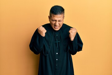 Young latin priest man standing over yellow background excited for success with arms raised and...