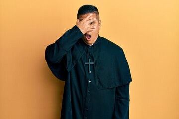 Young latin priest man standing over yellow background peeking in shock covering face and eyes with...