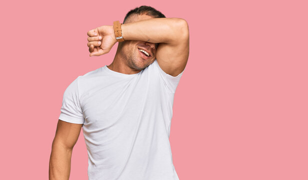 Handsome muscle man wearing casual white tshirt covering eyes with arm smiling cheerful and funny. blind concept.