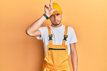 Hispanic young man wearing handyman uniform making fun of people with fingers on forehead doing loser gesture mocking and insulting.