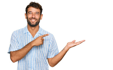 Handsome young man with beard wearing casual fresh shirt amazed and smiling to the camera while presenting with hand and pointing with finger.