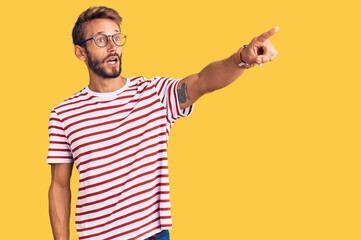 Fototapeta premium Handsome blond man with beard wearing casual clothes and glasses pointing with finger surprised ahead, open mouth amazed expression, something on the front