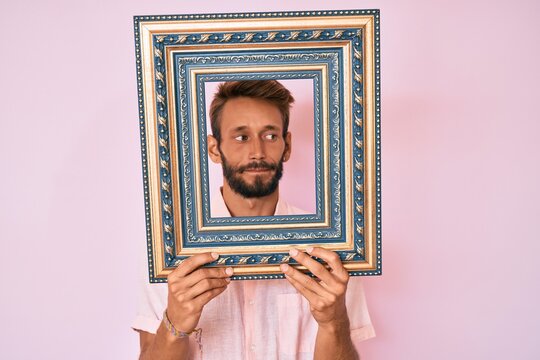 Handsome caucasian man with beard holding empty frame smiling looking to the side and staring away thinking.