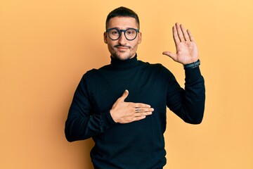 Handsome man with tattoos wearing turtleneck sweater and glasses swearing with hand on chest and...