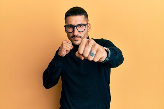 Handsome man with tattoos wearing turtleneck sweater and glasses punching fist to fight, aggressive and angry attack, threat and violence