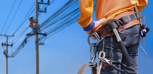 Rear view of electrician with safety belt and work tools is preparing to work on high altitude with blurred background of electrical workers team are working on power poles against blue sky