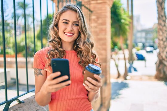 Young blonde girl smiling happy using smartphone and drinking take away coffee at the city.